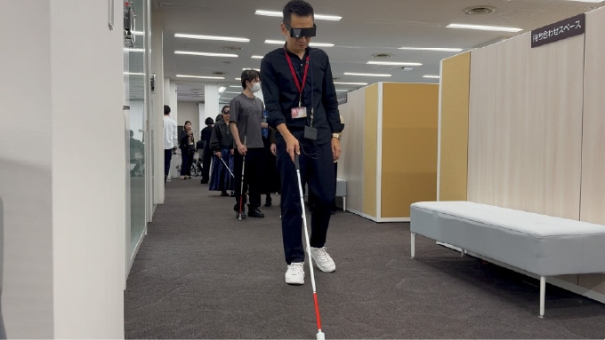 視覚障害体験の様子。ロービジョン体験メガネを着け、白杖を持った男性が通路を歩いている。