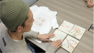 画像　リー イズミダ氏のアート作品と、そのアートをプリントしたTシャツを触り比べている様子　画像おわり