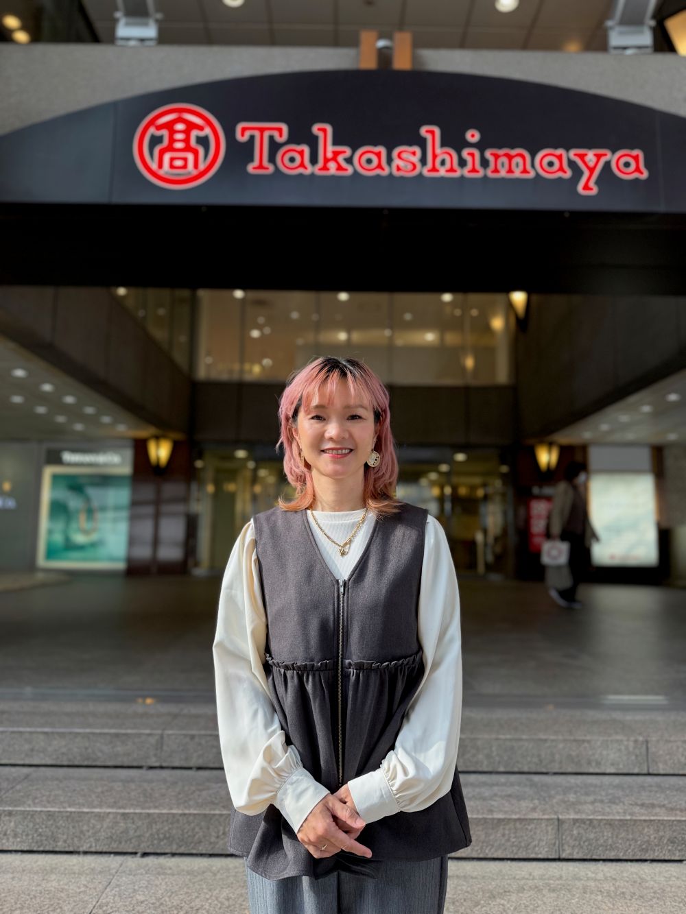 柏高島屋の正面入口前に立つピンクのショートヘアで白いブラウスにグレーのベストを重ね柔らかく微笑む宮田琴さん