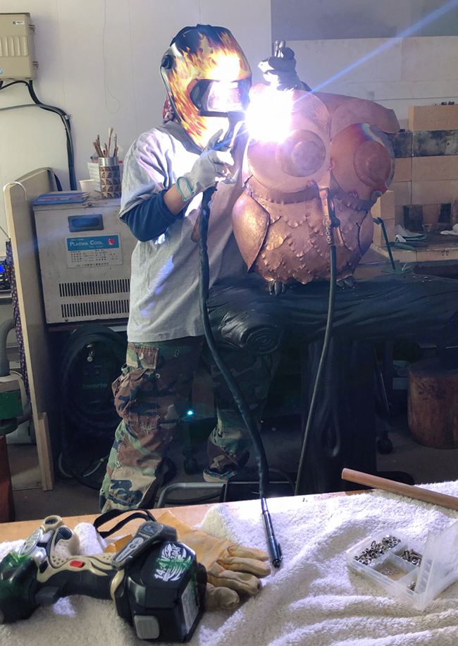 工房で溶接用のマスクを着けた宮田さんが強い光を放つ溶接の火花の前でふくろうの形をした銅色の立体作品に工具を当てている様子