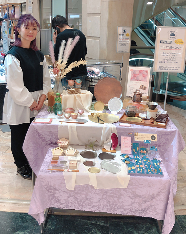 柏高島屋のPOP UP SHOPでピンク色の布にぐい呑みや箸置きを並べその横で優しく微笑む宮田琴さん