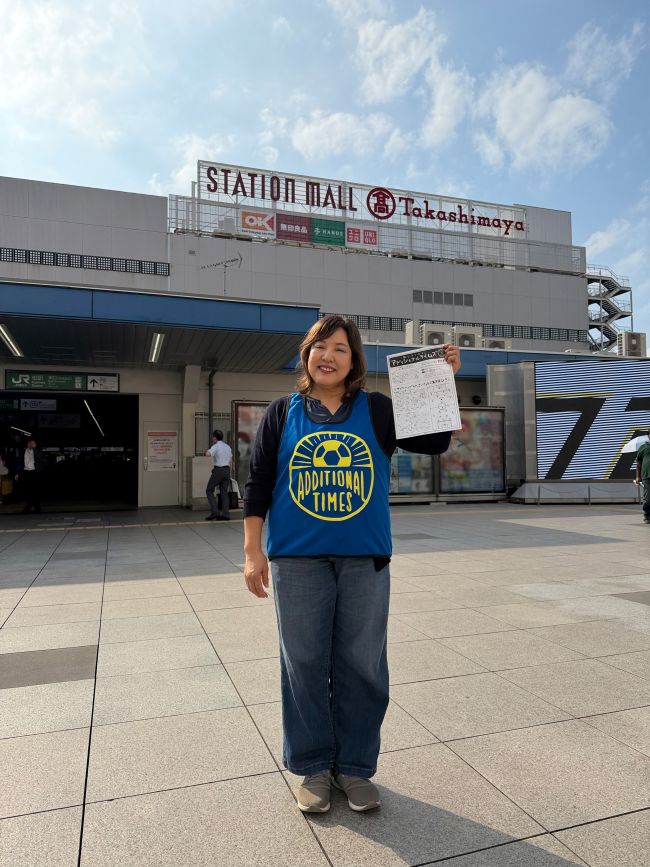 柏駅を背にアディショナルタイムズと書かれた青いビブスを着用しフリーペーパーを持つ加藤さん