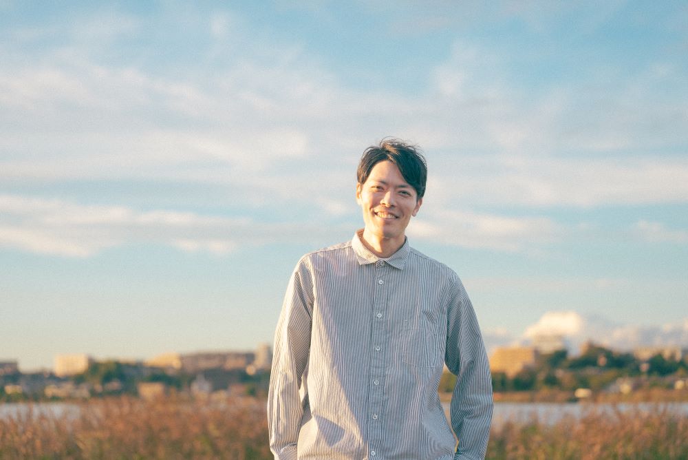 青空と雲の下で河川敷のような場所に立ち穏やかに微笑む藤田さん