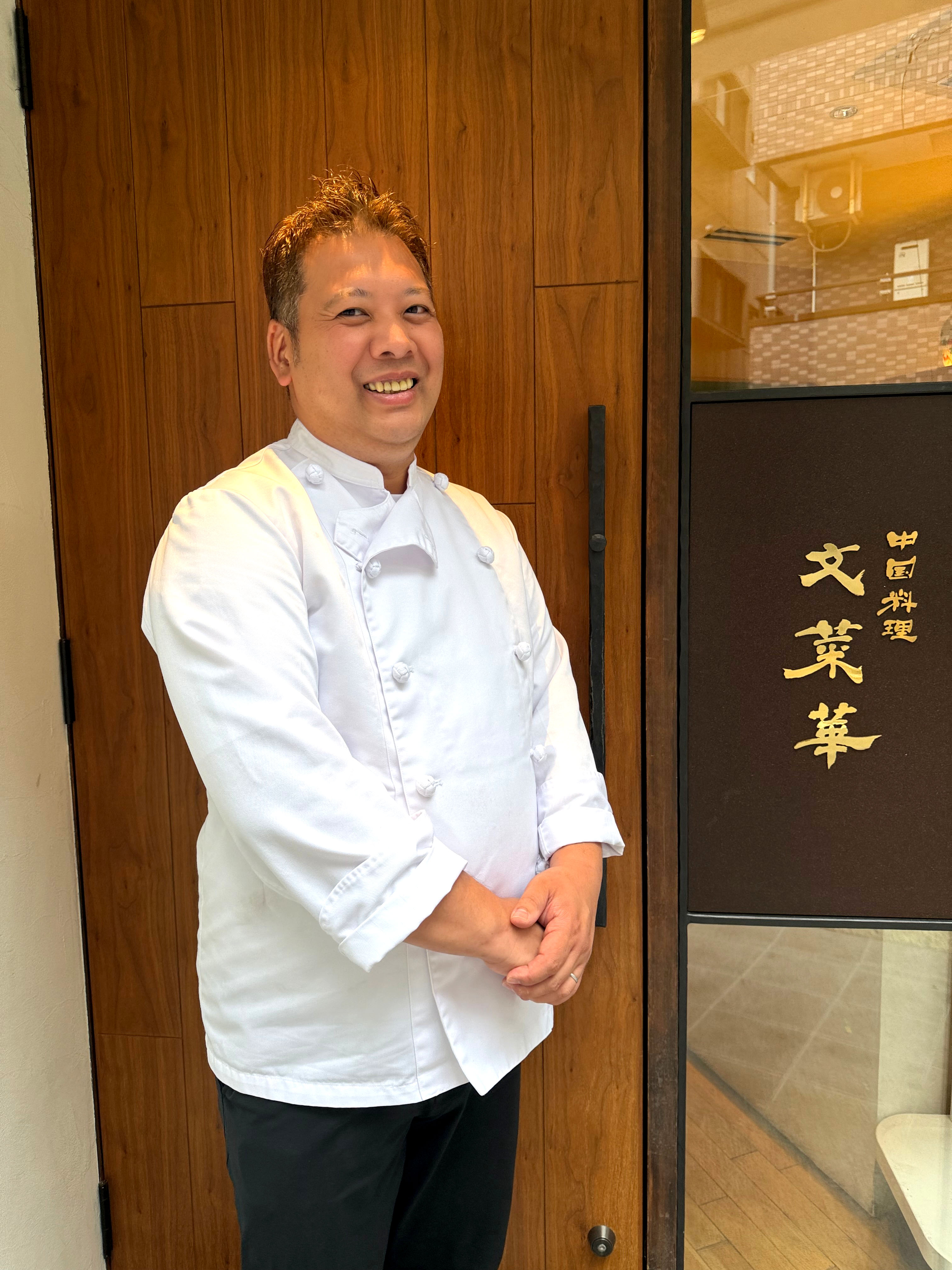 中国料理 文菜華の入口前で白いコックコート姿で笑顔を見せる渡辺シェフ