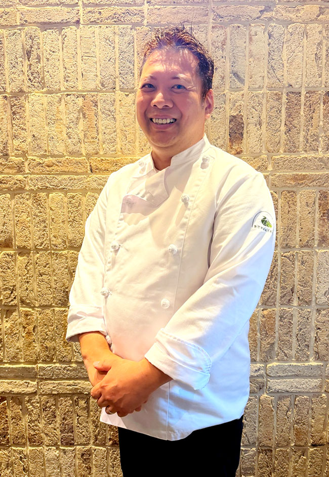 中国料理 文菜華の店内で石壁を背景に立つ渡辺シェフ