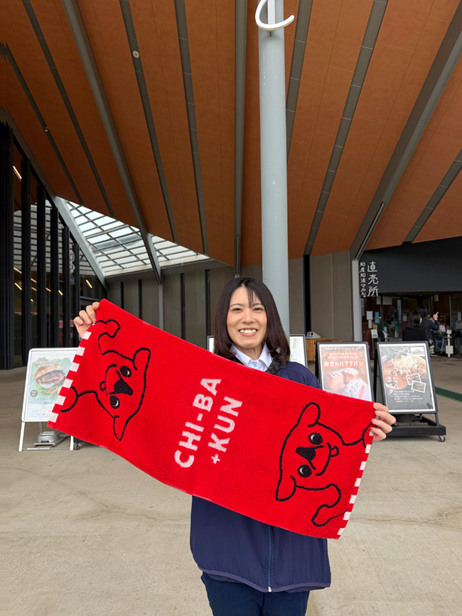チーバくんの赤いタオルを見せながら道の駅しょうなんの建物の前に笑顔で立つ木村さん